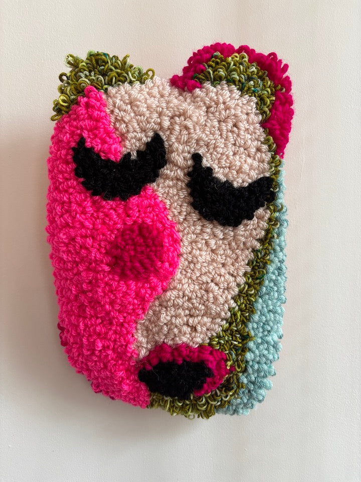 Colorful yarn art piece of a face with pink cheeks, black eyes, and green hair on a white background.